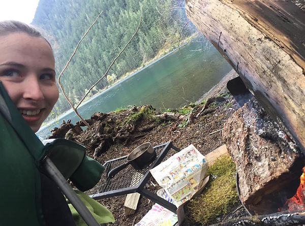 Aubrey Frei outdoors beside a river, smiling, with drawing supplies and a campfire nearby